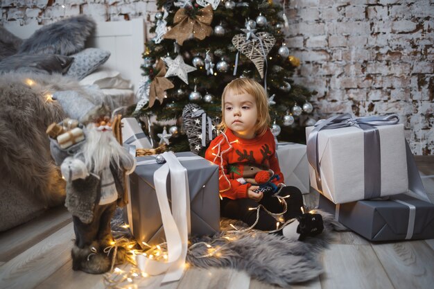 Une petite fille vêtue d'un pull rouge chaud est assise sous un sapin de Noël avec des jouets et des cadeaux. Enfance heureuse. Ambiance de vacances du nouvel an