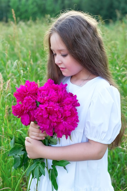 Une petite fille vêtue d'une robe blanche avec un bouquet de pivoines roses sur un fond vert flou Séance photo dans la nature