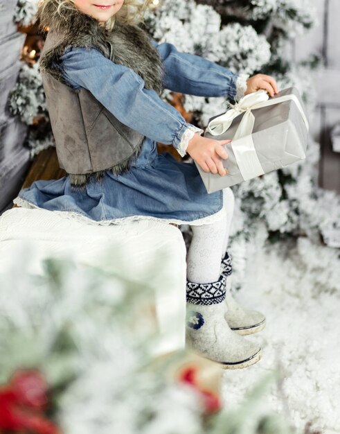 Une petite fille vêtue d'une robe bleue, d'une veste sans manches et de collants blancs est assise dans un décor du Nouvel An sur un banc avec un cadeau du Nouvel An dans les mains. photo