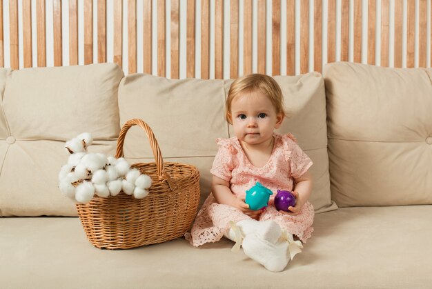 Petite fille vêtue d'une robe est assise et regarde la caméra sur un canapé beige avec un panier et des œufs colorés