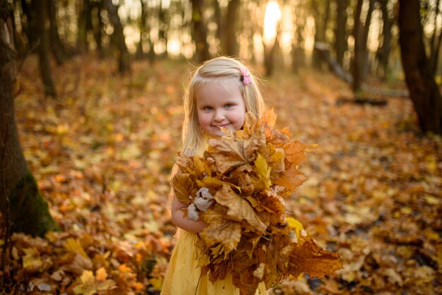 Petite fille vêtue d'une robe jaune dans le parc en automne. enfant tient un tas de feuilles dans ses mains. Fermer.