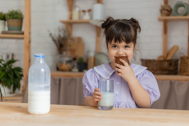 une petite fille vêtue d'une robe lilas boit du lait et mange un petit pain à la table de la cuisine. espace pour le texte, bannière