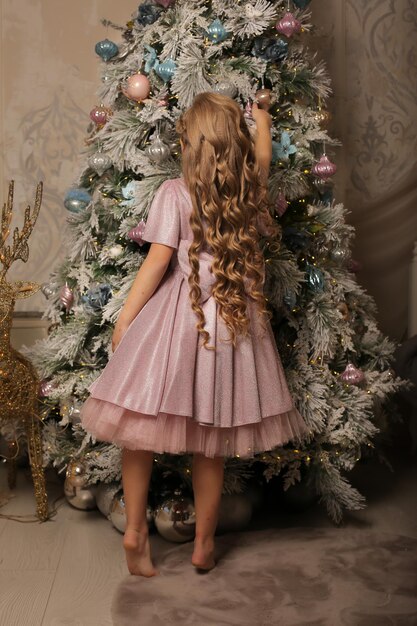 une petite fille vêtue d'une robe rose décore un arbre de Noël avec des jouets