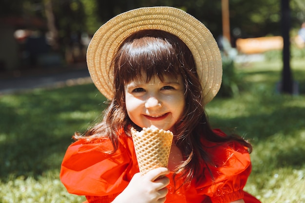 Une petite fille vêtue d'une robe rouge et d'un chapeau de paille mange de la glace dans un cône du parc