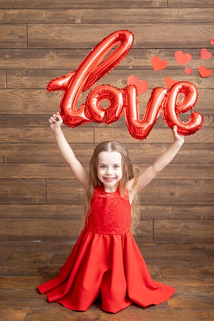 Une petite fille vêtue d'une robe rouge tient un grand ballon avec l'inscription Love sur un fond en bois brun foncé avec des coeurs rouges le concept de la Saint-Valentin une place pour le texte