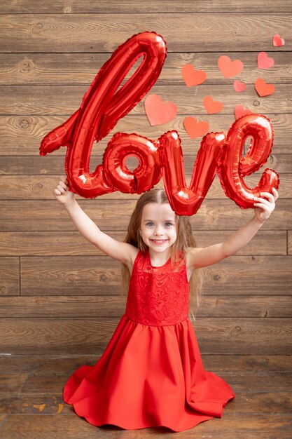 Une petite fille vêtue d'une robe rouge tient un grand ballon avec l'inscription Love sur un fond en bois brun foncé avec des coeurs rouges le concept de la Saint-Valentin une place pour le texte