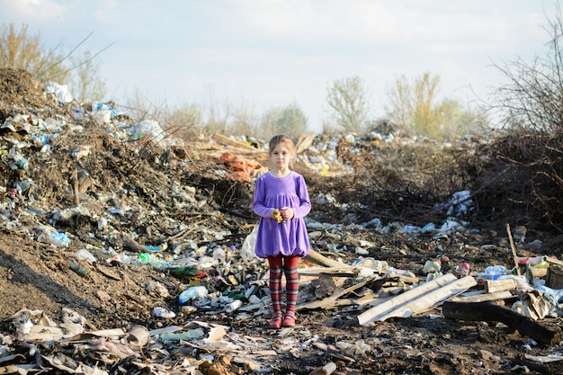 Petite fille vêtue d'une robe violette et de collants à rayures rouges dans un dépotoir parmi des piles d'ordures contenant des fleurs fanées jaunes