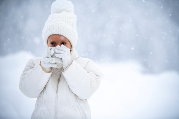 Une petite fille vêtue d'une veste blanche et d'un chapeau blanc tient une tasse de thé dans ses mains lors d'une chute de neige sur fond de belles congères et de forêts