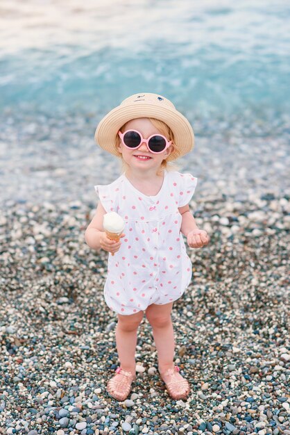 Une petite fille vêtue de vêtements d'été, d'un chapeau jaune et de lunettes de soleil roses se tient sur la plage, mange de la glace blanche et regarde la caméra.