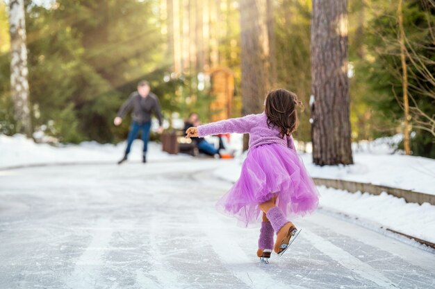 Petite jeune fille vêtue d'un pull rose et d'une jupe ample se promène par une journée d'hiver ensoleillée sur une patinoire extérieure dans le parc