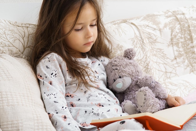 Petite jolie adorable jolie fille brune en pyjama lisant un livre avec un ours en peluche sur un lit moelleux Apprendre à l'éducation à domicile