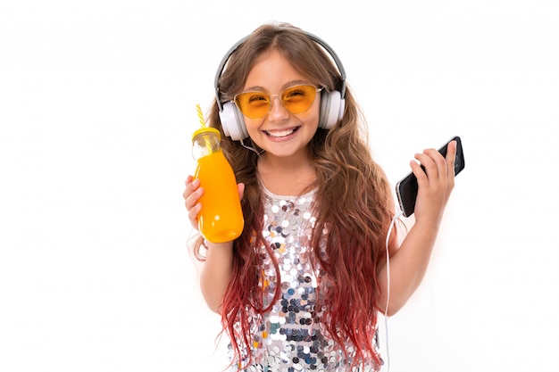 Petite jolie fille caucasienne écouter de la musique avec de gros écouteurs et boit du jus d'orange, photo isolé sur fond blanc