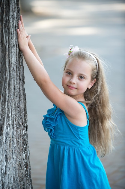 Petite petite fille ou enfant heureux mignon avec un adorable visage souriant et un arc dans les cheveux blonds en robe bleue en été en plein air près de l'arbre sur fond flou