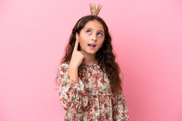 Petite princesse caucasienne avec couronne isolée sur fond rose pensant une idée pointant le doigt vers le haut