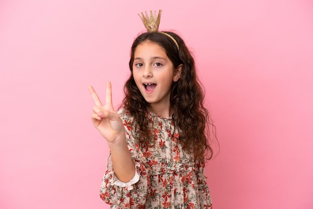 Petite princesse caucasienne avec couronne isolée sur fond rose souriant et montrant le signe de la victoire