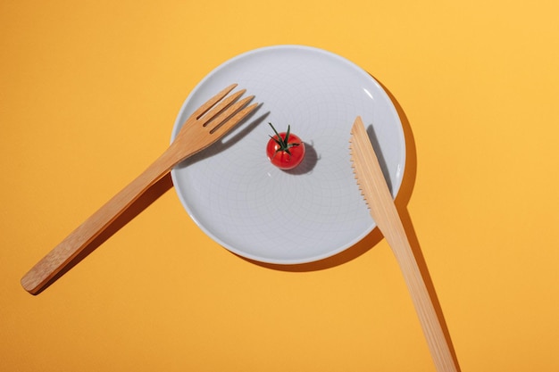 Une petite tomate cerise rouge sur une assiette et une fourchette et un couteau en bambou sur fond jaune