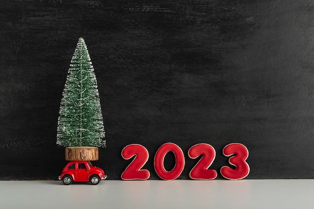 La petite voiture porte un petit sapin de Noël sur fond d'inscription 2023 Nouvel An arrive Fond sombre