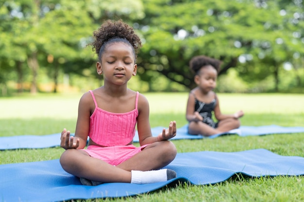 Petites filles afro-américaines les yeux fermés assis sur un tapis roulant pratiquent la méditation yoga dans le parc