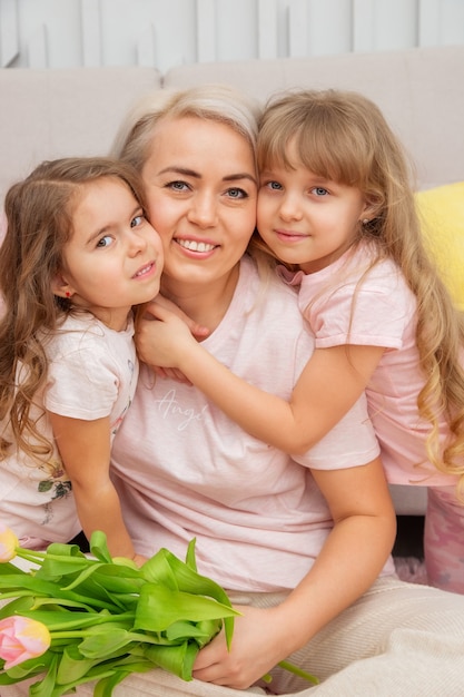 De petites filles d'apparence caucasienne se serrent dans leurs bras avec leur mère, assise sur le sol devant le canapé dans un salon lumineux dans un style scandinave