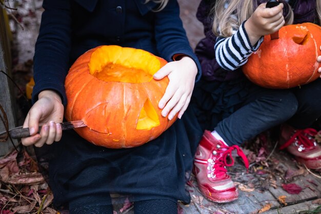 Les petites filles fabriquent une citrouille-lanterne à partir de grosses citrouilles pour célébrer les vacances d'halloweenManteau de chapeau de costume de sorcière Couper avec un couteauEnlever la pulpe avec des grainesActivité de plein air arrière-courFête des enfants