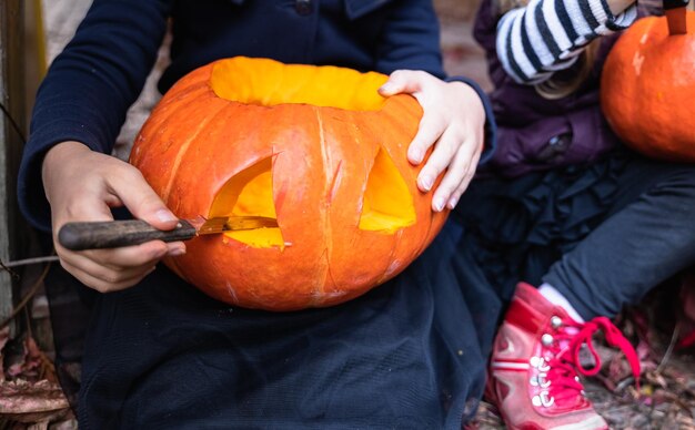 Les petites filles fabriquent une citrouille-lanterne à partir de grosses citrouilles pour célébrer les vacances d'halloweenManteau de chapeau de costume de sorcière Couper avec un couteauEnlever la pulpe avec des grainesActivité de plein air arrière-courFête des enfants