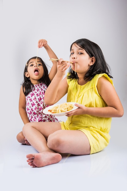 Petites filles ludiques indiennes ou asiatiques mignonnes mangeant de délicieuses nouilles chinoises avec une fourchette ou des baguettes, isolées sur fond coloré