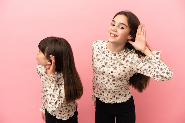 Petites filles sœurs isolées sur fond rose en écoutant quelque chose en mettant la main sur l'oreille