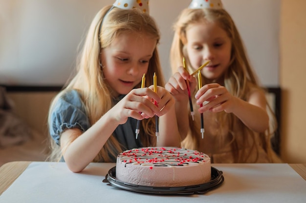 Les petites filles soeurs jumelles sont heureuses de décorer le gâteau d'anniversaire avec des bougies chapeau de vacances fait maison fête d'anniversaire de l'enfant fête à l'intérieur pendant l'isolement de la quarantaine de maladie