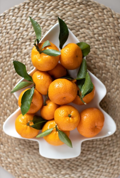 Photo petites mandarines mûres avec des feuilles, sur une assiette en forme d'arbre de noël sur une serviette de service en osier. concept d'aliments vitaminés d'hiver. composition festive de moody nature morte. pour carte de voeux, livre, histoires.