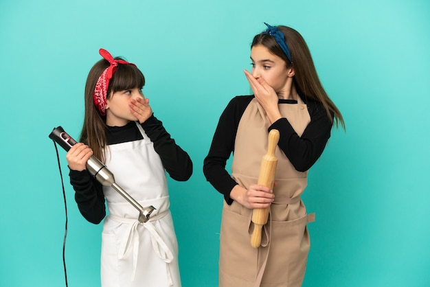 Petites soeurs cuisinant à la maison isolées sur fond bleu couvrant la bouche avec les mains pour avoir dit quelque chose d'inapproprié