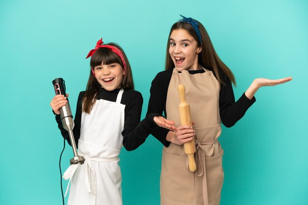 Petites soeurs cuisinant à la maison isolées sur fond bleu avec une expression faciale surprise et choquée