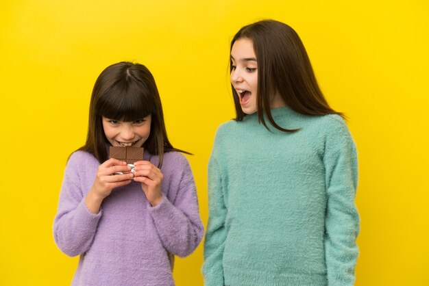 Petites sœurs isolées sur fond jaune mangeant une tablette de chocolat