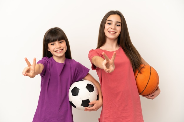 Petites sœurs jouant au football et au basket-ball isolé sur fond blanc souriant et montrant le signe de la victoire