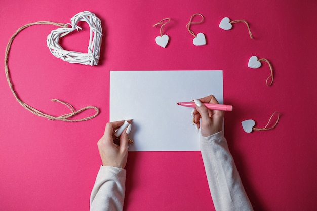 Petits coeurs en bois blanc et grand coeur en bois en osier blanc avec une corde sur fond rose avec copie espace. Carte de Saint Valentin avec des mains de femme écrivant avec un stylo rose sur une feuille de papier blanc.