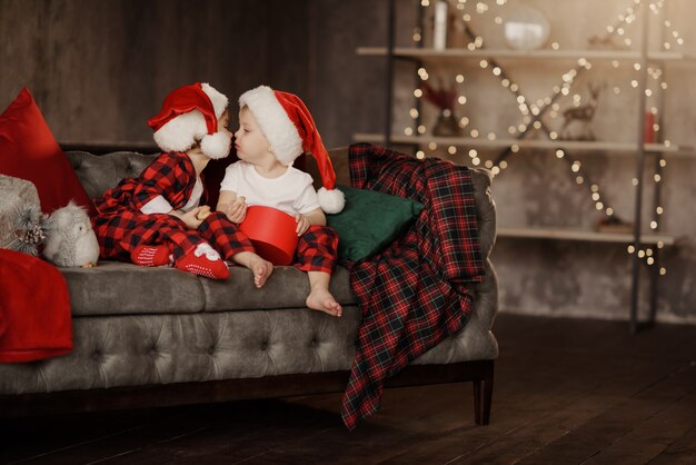 Petits enfants mignons en chapeaux de Père Noël assis sur une grande chaise confortable dans la chambre avec boîte de cadeaux de Noël