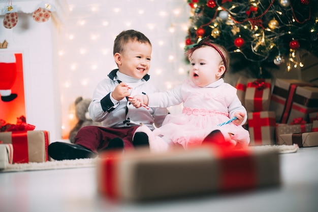 Petits enfants près de l'arbre de Noël et de la cheminée avec des bonbons colorés dans les mains