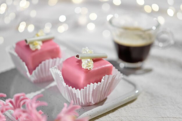 Petits fours de la Saint-Valentin avec glaçage au massepain et fleurs à la crème. Café expresso dans une tasse en verre. Joyeuse saint Valentin! Guirlande de lumières sur nappe textile ivoire, blanc cassé. Fleur de jacinthe rose.