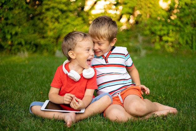 Les petits garçons dans les écouteurs sans fil tiennent un ordinateur tablette à l'extérieur dans un parc sourire communiquent