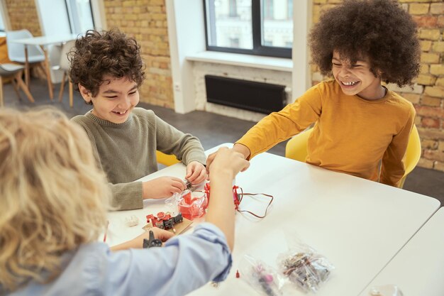 Petits garçons divers et positifs riant assis à la table et examinant les détails d'un robot