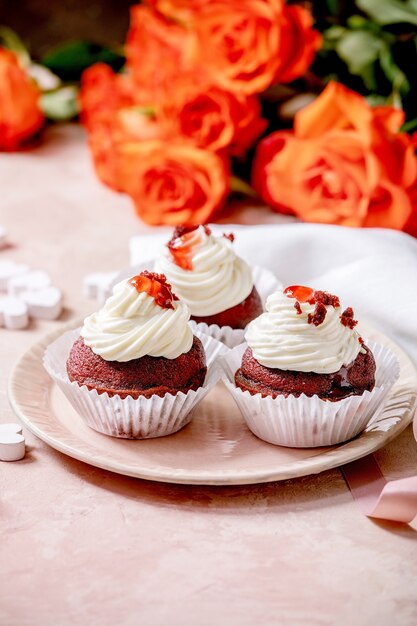 Petits gâteaux de velours rouge faits maison avec de la crème fouettée sur plaque en céramique rose, serviette blanche avec ruban, fleurs roses, coeurs en bois sur mur de texture rose