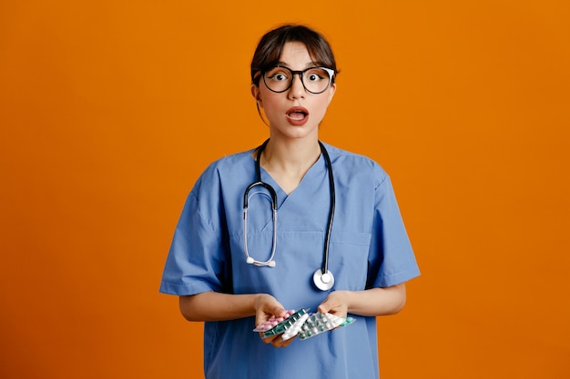 Peur tenant des pilules jeune femme médecin portant un stéthoscope fith uniforme isolé sur fond orange