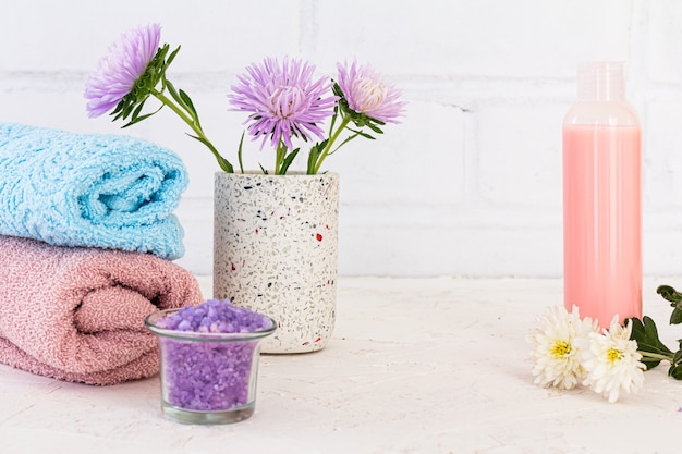 Peut avec du sel de mer, une bouteille de shampoing, des serviettes et des fleurs d'asters sur une surface blanche