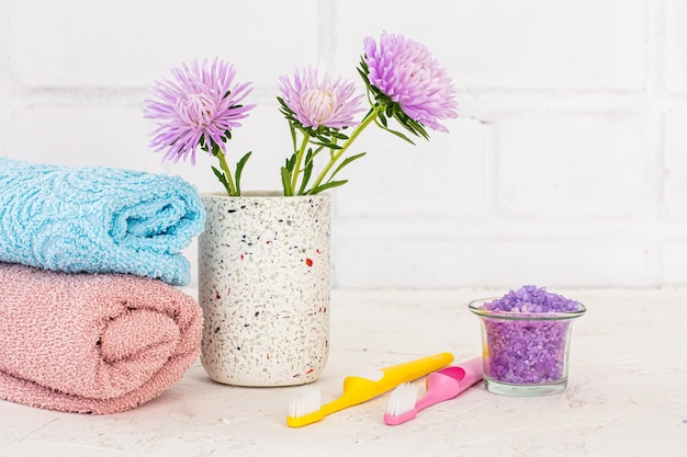 Peut avec du sel de mer, des serviettes, des brosses à dents et des fleurs d'asters sur fond blanc. Cosmétiques femme et accessoires de lavage.