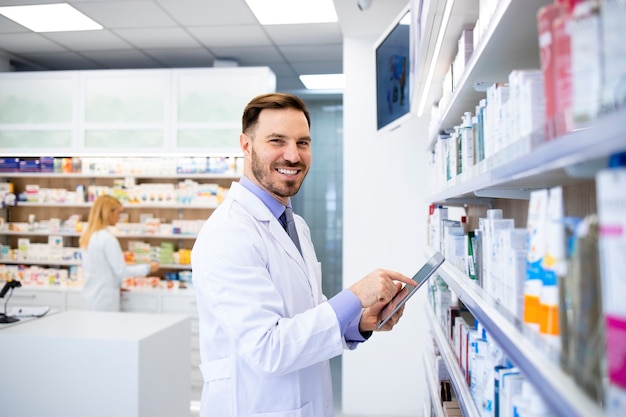 Pharmacien debout près des étagères avec des médicaments et en tapant sur tablette en pharmacie.