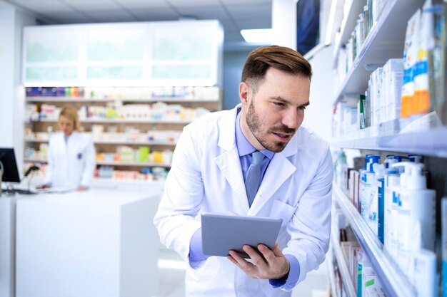 Pharmacien professionnel beau mâle en blouse blanche tenant la tablette et regardant les médicaments en pharmacie ou en pharmacie.