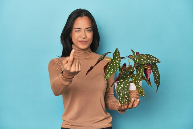 Philippine tenant une plante sur le studio bleu pointant avec le doigt sur vous comme si invitant venir plus près