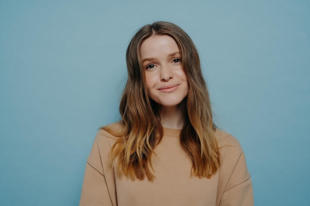 Photo d'une adolescente assez souriante sans maquillage regardant la caméra portant un pull marron clair confortable posant isolée sur fond bleu bleu studio. Notion de jeunes