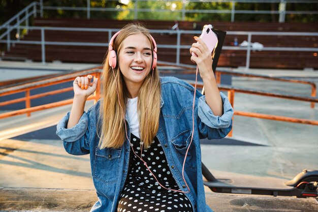 Photo d'une adolescente souriante et positive à l'extérieur dans un parc, écoutant de la musique avec des écouteurs tenant un téléphone portable.