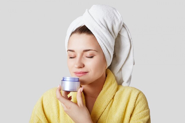 Photo De L'adorable Jeune Femme Heureuse Détient Une Bouteille De Crème Et La Sent, Va Faire Un Masque Sur Le Visage Après Avoir Pris Un Bain, Isolé Sur Le Mur Blanc. Concept De Personnes, De Cosmétiques Et De Beauté.