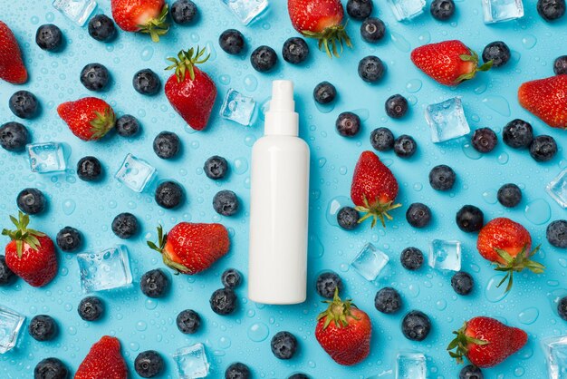 Photo aérienne de fraises bleuets partout gouttes d'eau glaçons et bouteille blanche de spray au milieu isolé sur fond bleu
